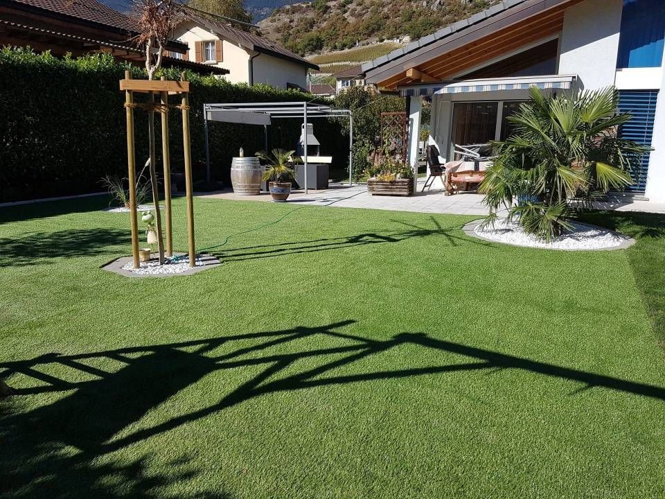 Cour arrière ensoleillée avec pelouse verte, patio et maison, agrémentée de plantes en pot et d'une structure.