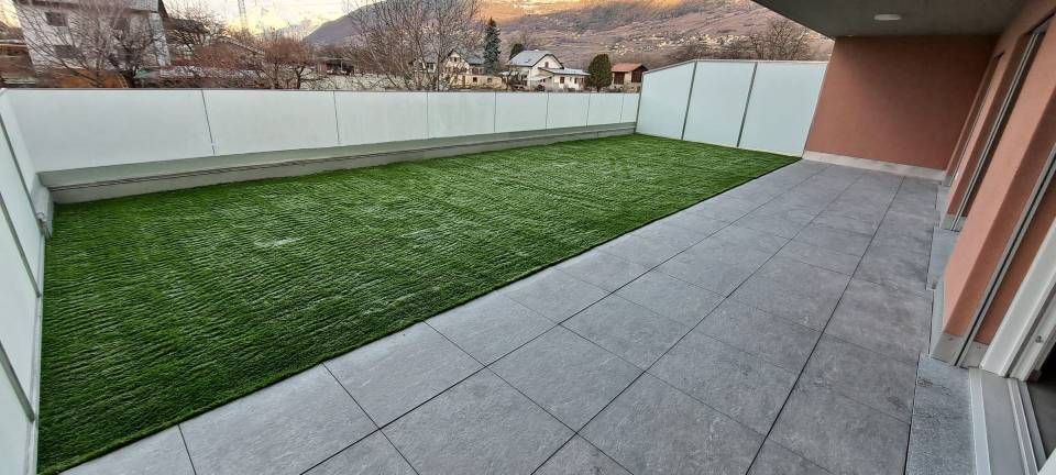 Terrasse moderne dans le jardin avec pavés gris, gazon artificiel, clôture blanche et vue sur les collines.