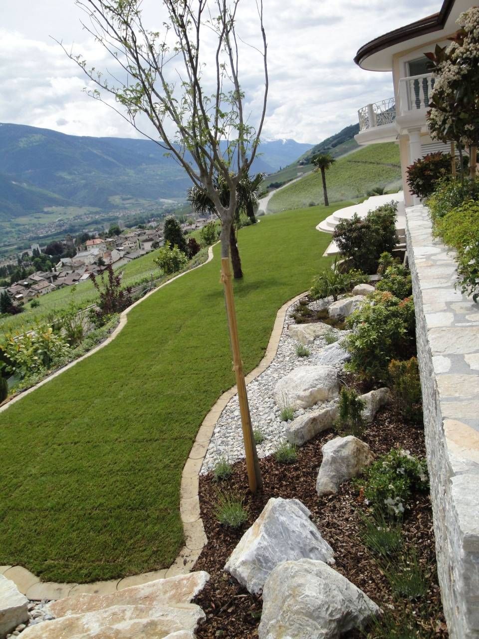 Jardin en terrasses à flanc de colline, bordé de pierres, avec un jeune arbre et une vue sur la vallée, à côté d'une maison.