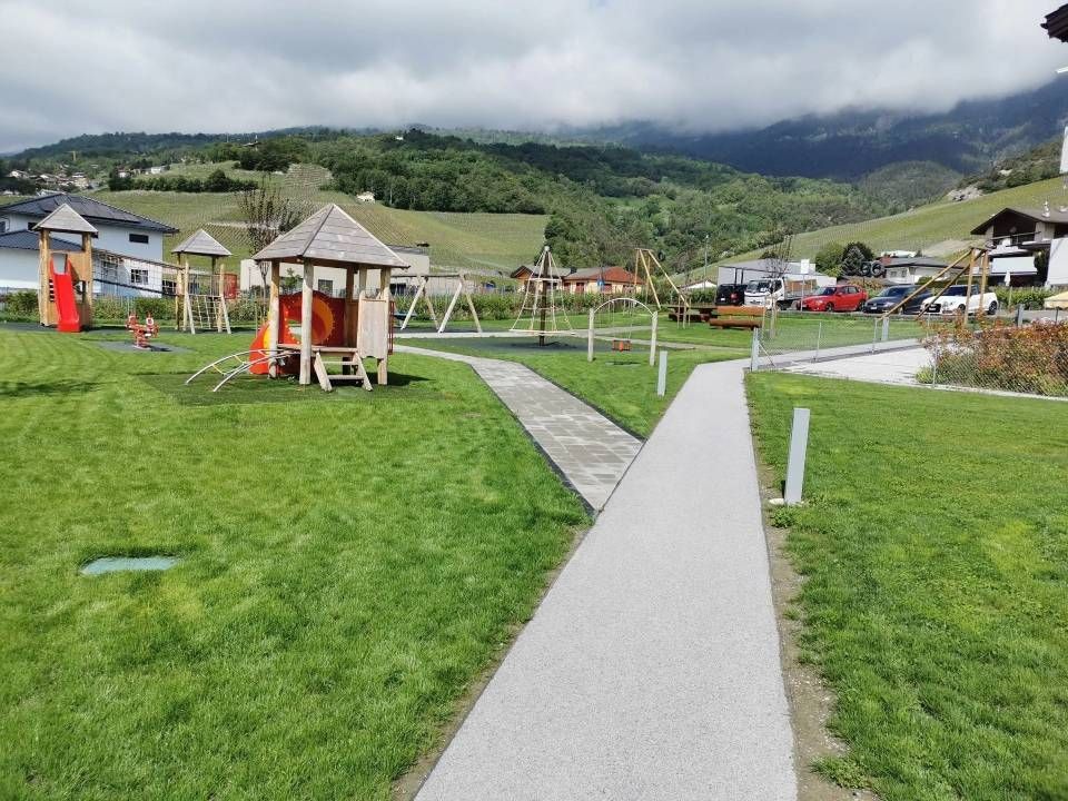 Parc nuageux avec des pelouses vertes, un chemin pavé et de petits kiosques donnant sur les collines et les maisons.