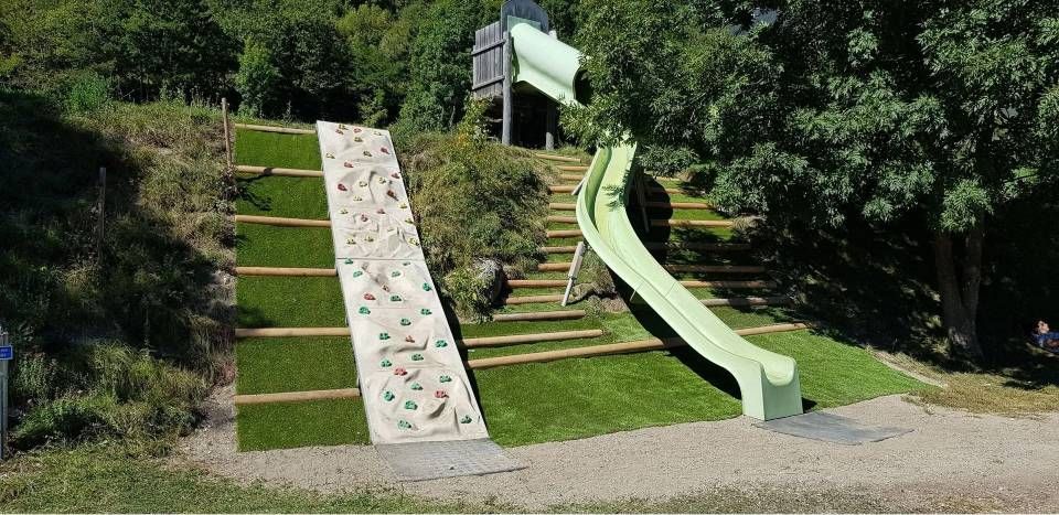 Aire de jeux extérieure avec un mur d'escalade et un toboggan vert sur une colline herbeuse.