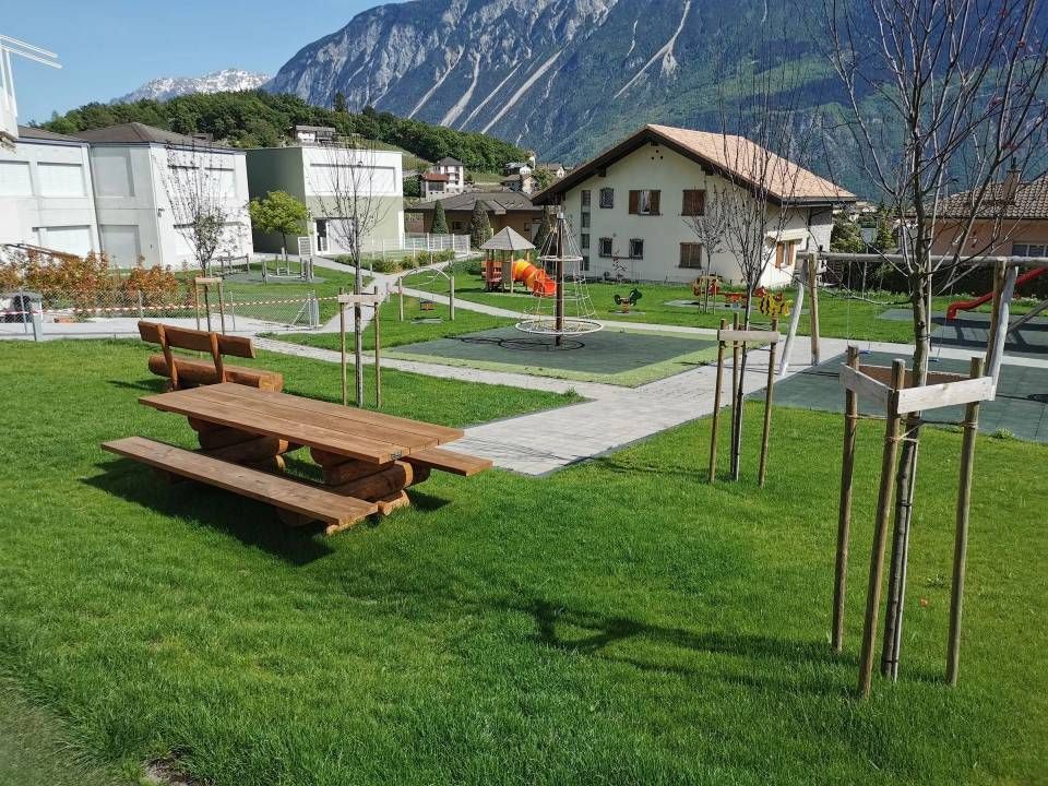 Parc avec banc en bois, pelouse, allées, jeunes arbres, maisons et montagnes en arrière-plan.