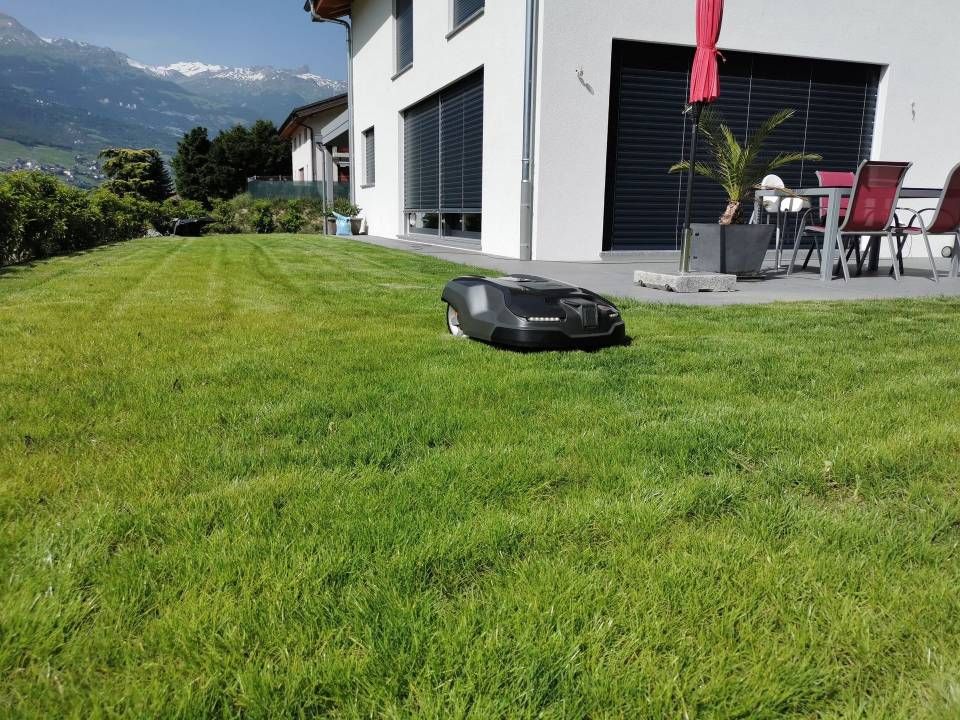 Tondeuse robotisée sur une pelouse verte à côté d'une maison moderne avec des montagnes en arrière-plan