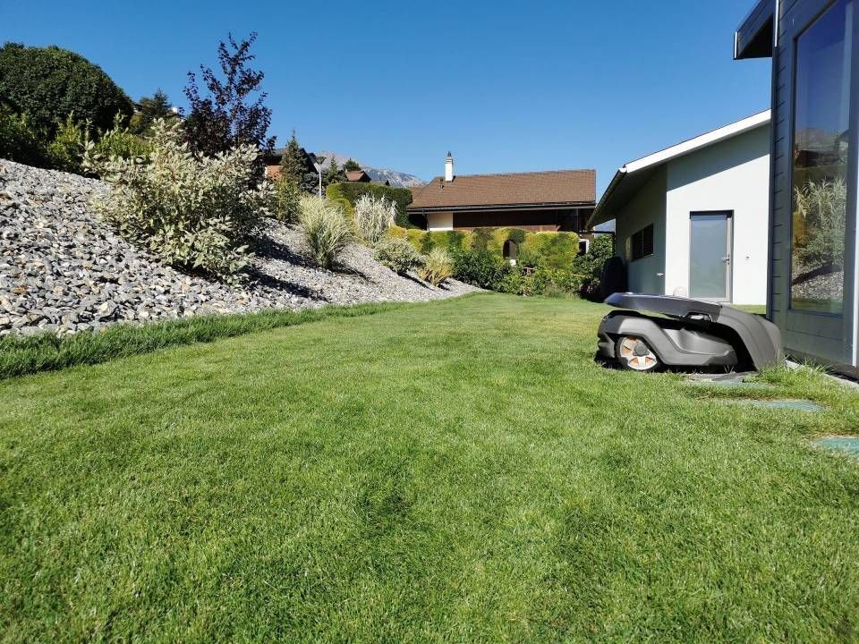 Cour arrière gazonnée jouxtant une maison moderne, avec une pente en pierre et une tondeuse robotisée près du garage.