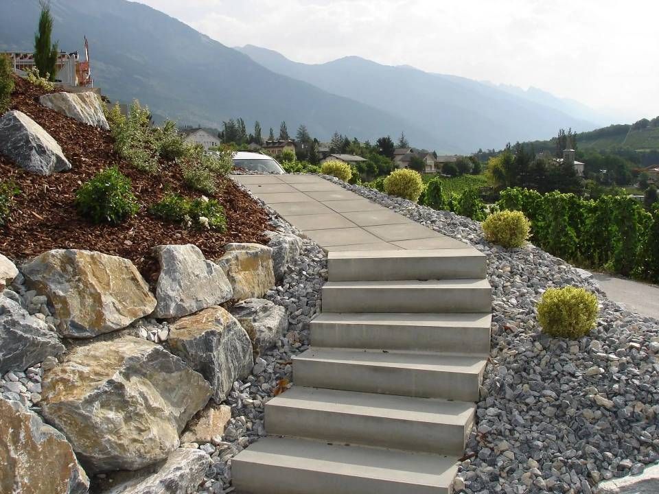 Des marches en pierre traversent un jardin de gravier et mènent à flanc de colline, avec les montagnes en arrière-plan.