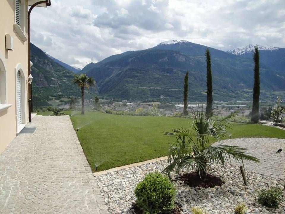 Jardin de la villa avec allée de pierre, pelouse verte, cyprès et vue sur la montagne sous un ciel nuageux