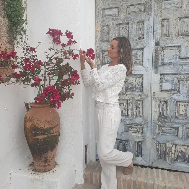 Una mujer vestida de blanco de pie junto a un jarrón de flores rojas.