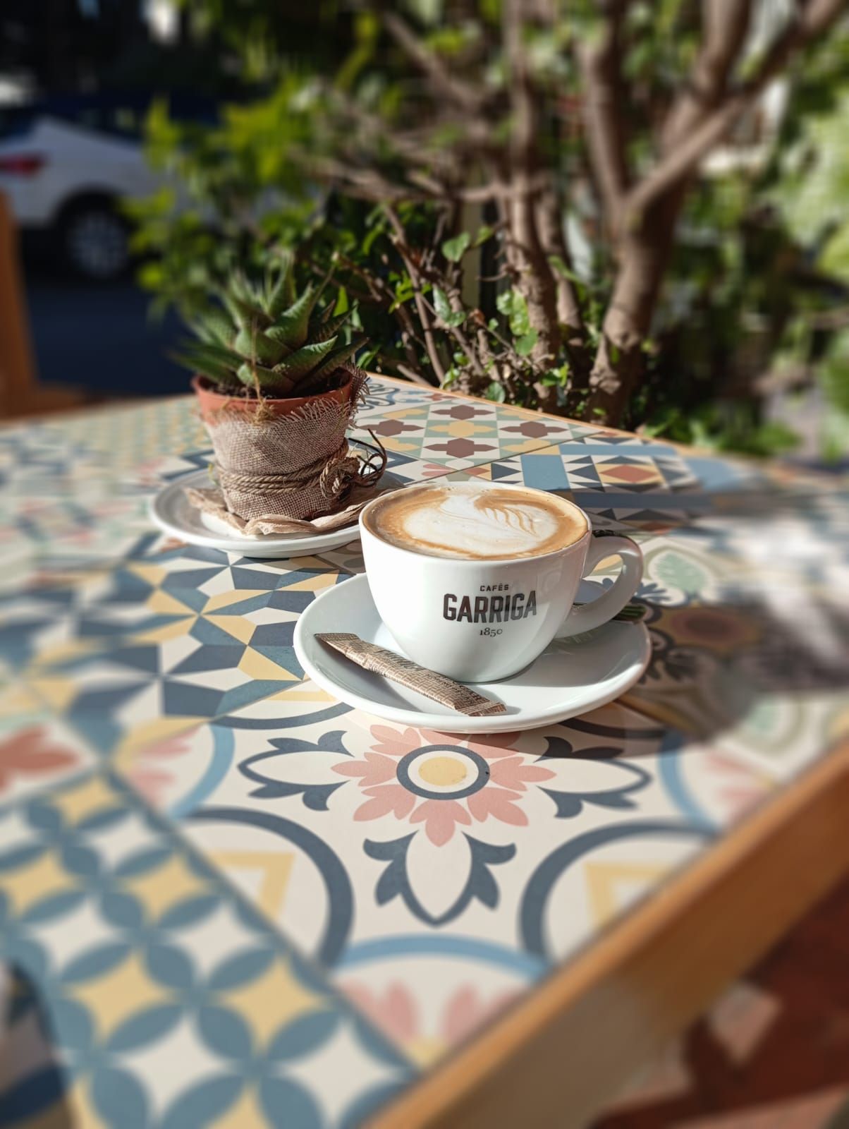 Taza de café con arte latte sobre una mesa estampada, una pequeña planta y un árbol en el fondo.
