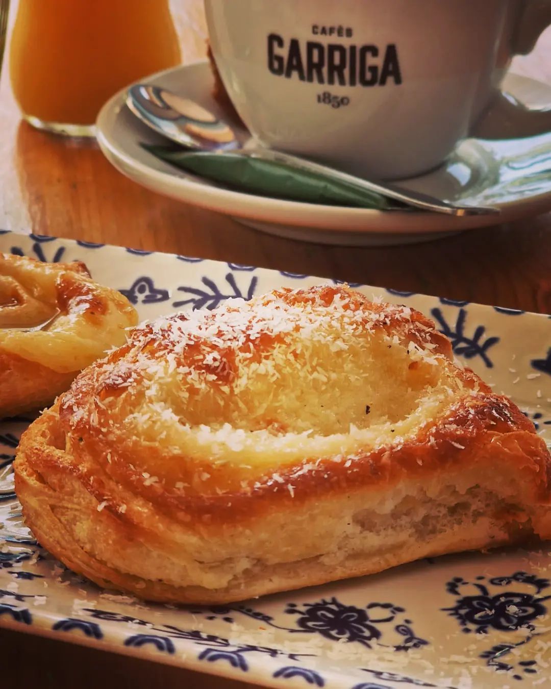 Pastel con coco rallado sobre plato estampado, taza de café y zumo de naranja.