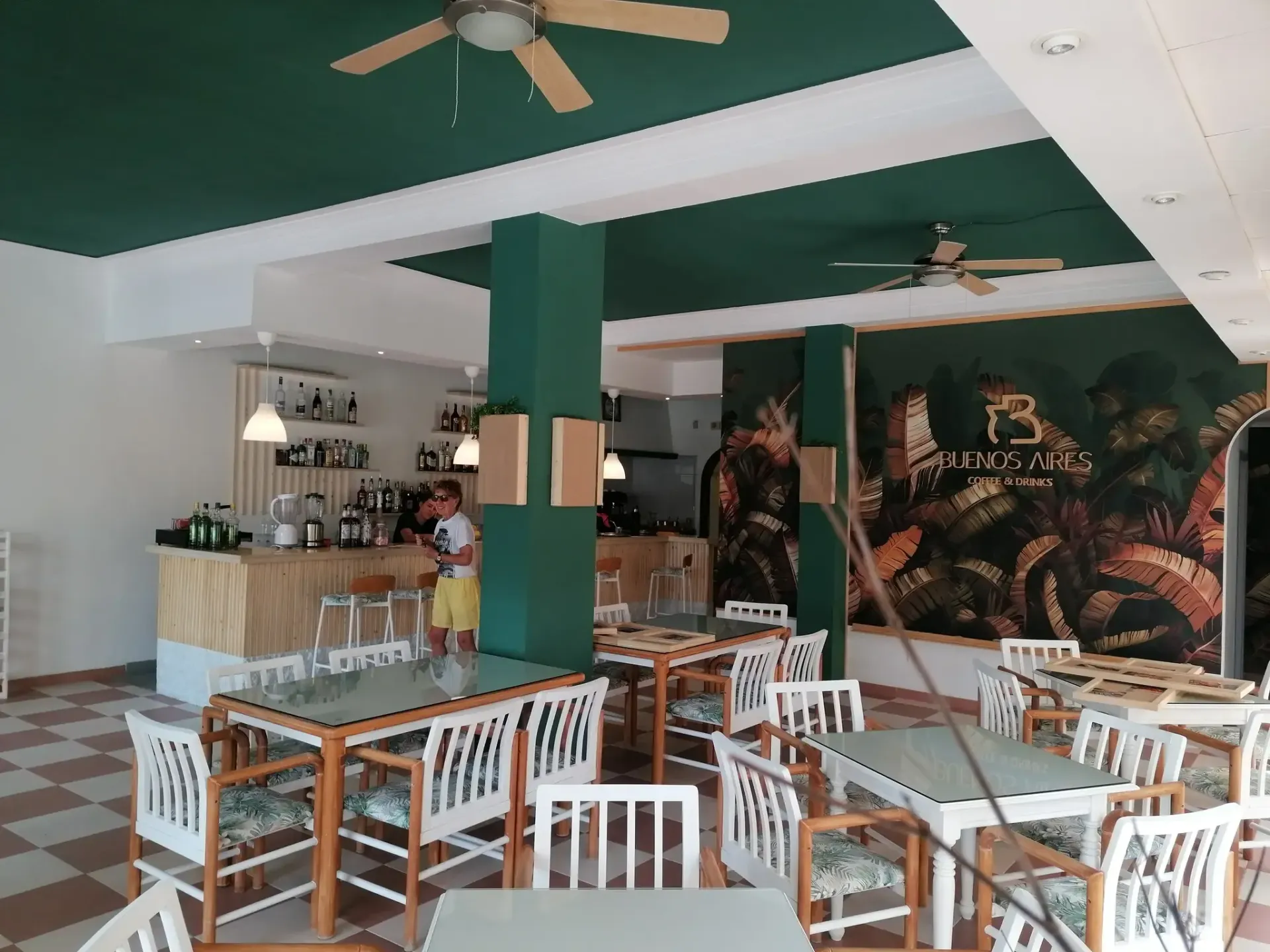 Interior de una cafetería con mesas y sillas, una barra, techo verde y un mosaico en la pared.