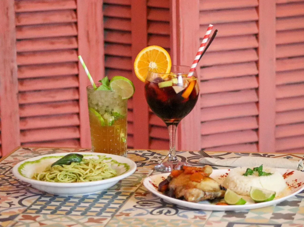 Pasta, arroz con carne y bebidas sobre una mesa con fondo de persianas rosas.