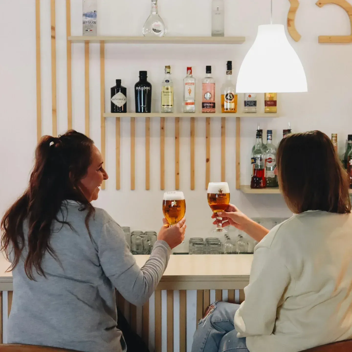 Dos mujeres brindan con cervezas en un bar. Estantes de madera con botellas de licor al fondo.