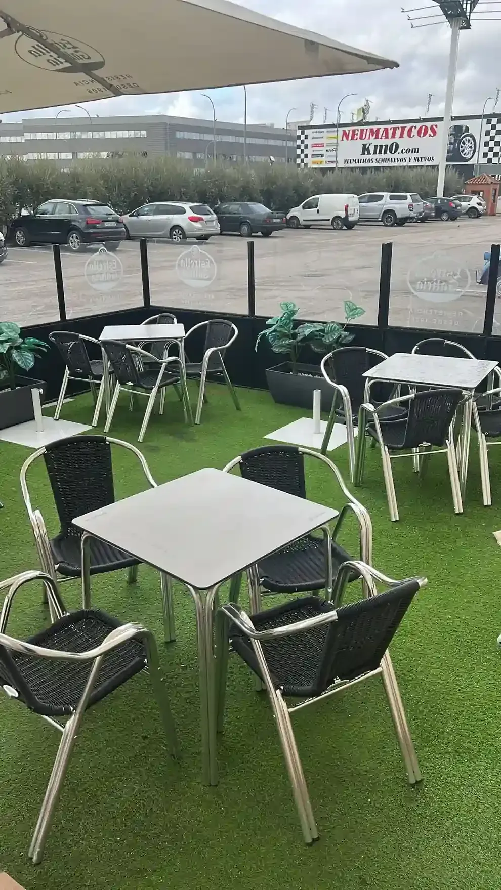 Zona de estar tipo cafetería al aire libre con mesas y sillas sobre césped artificial, cielo nublado.