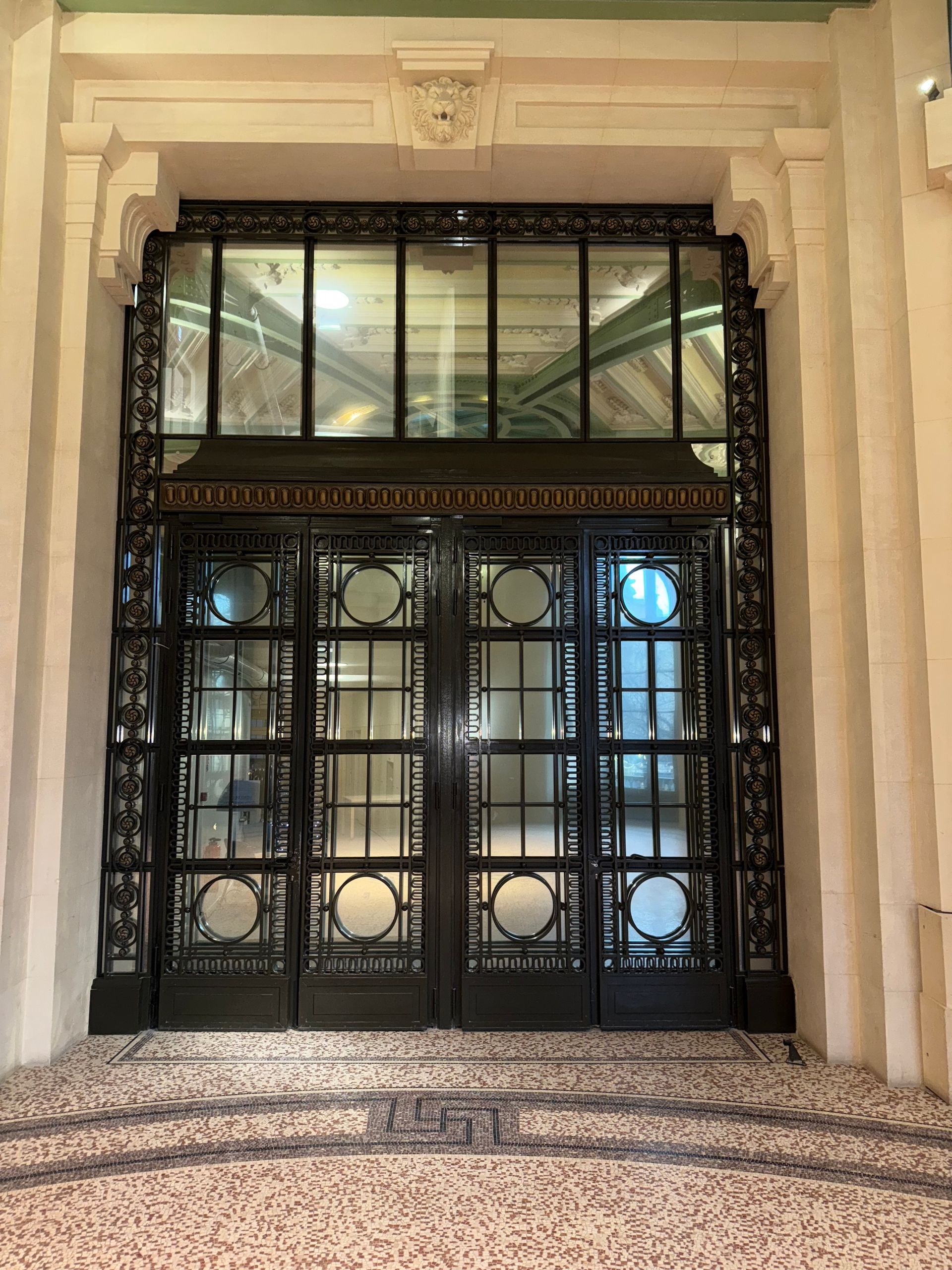 Double porte en fer forgé noir ouvragé, avec panneaux de verre et imposte, intégrée dans un hall d'entrée en pierre architecturale.