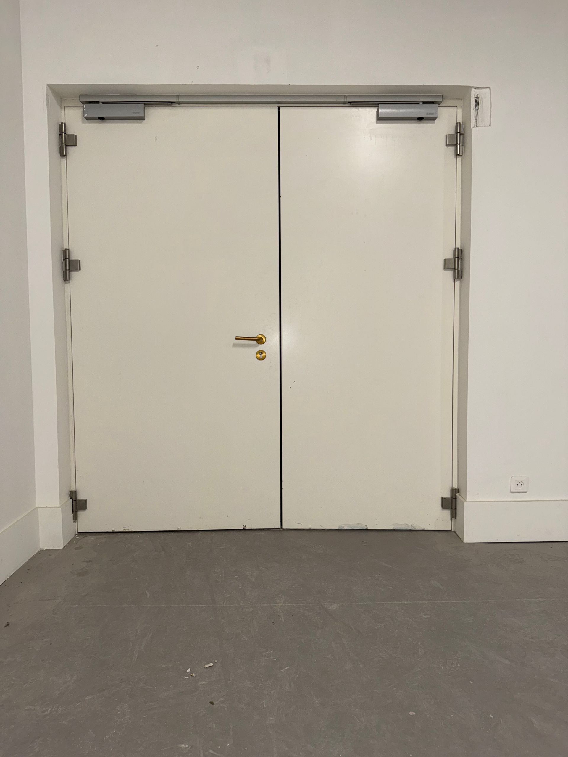 Deux portes doubles fermées, blanc cassé, avec des ferrures et des charnières argentées, dans un couloir intérieur blanc uni.