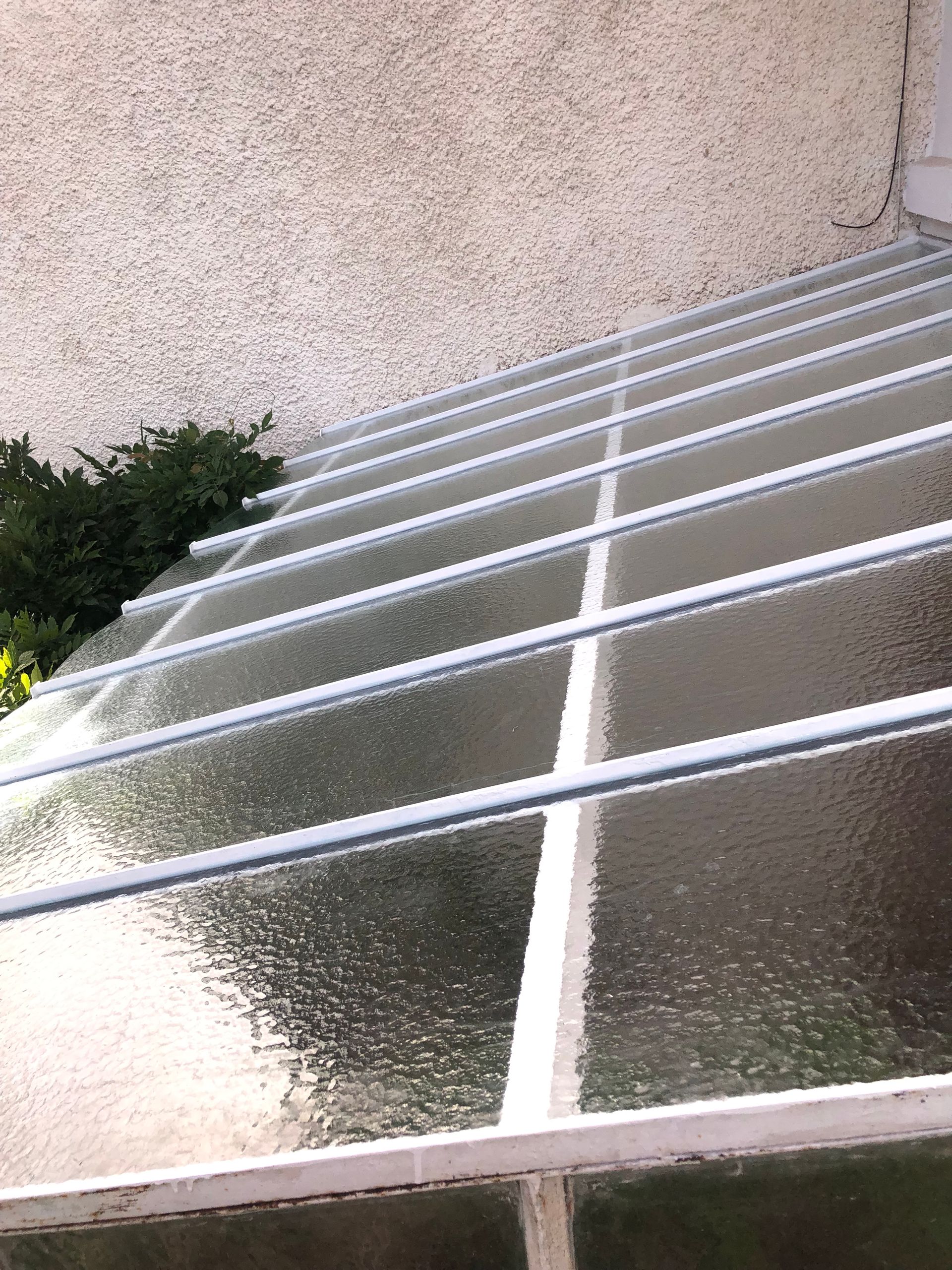 Vue en biais d'un toit en appentis en verre translucide texturé, avec des poutres de soutien en métal blanc, contre un mur en crépi.