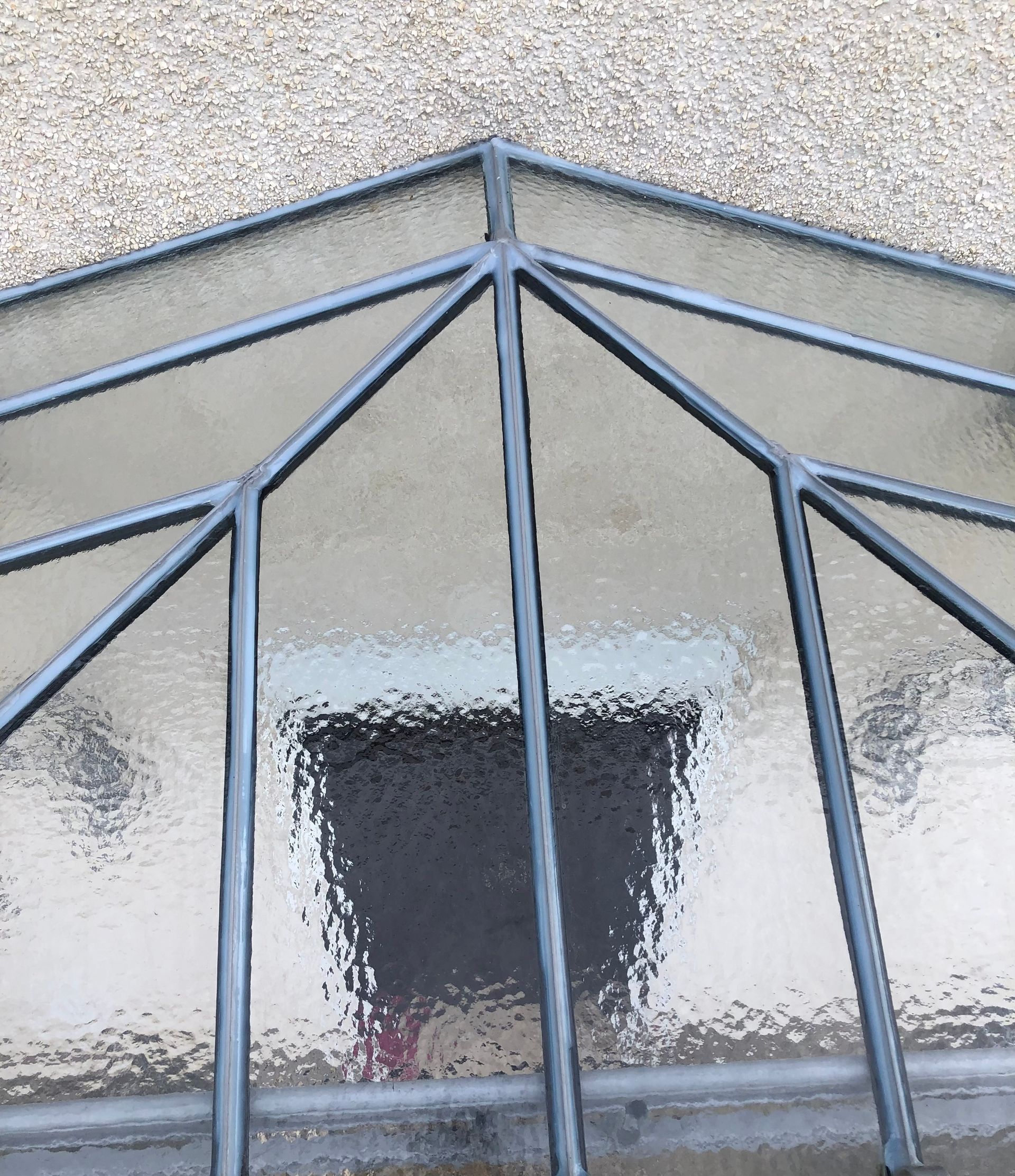 Vue plongeante à travers une verrière géométrique à cadre sombre, révélant le reflet flou d'une porte sombre.