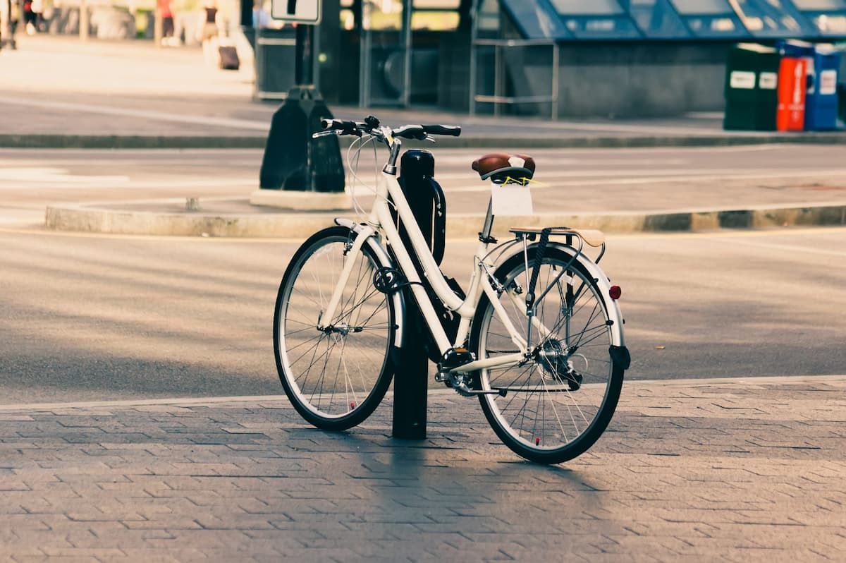 Una bicicleta blanca está atada con candado a un poste negro en una calle pavimentada de la ciudad, cerca de una acera.