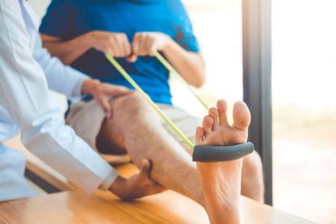 In einem hellen Studio hilft ein Physiotherapeut einem Patienten bei der Durchführung einer Beinübung mit einem gelben Widerstandsband.