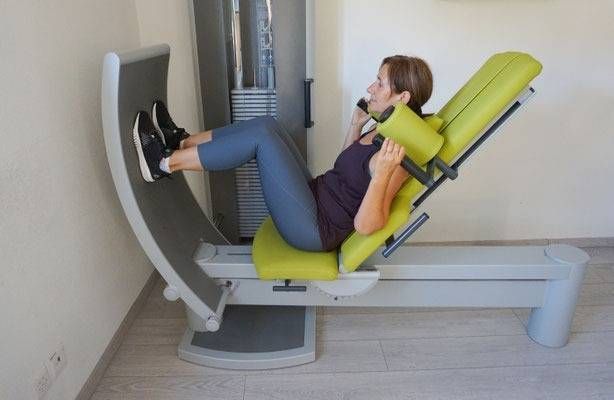 Eine Person benutzt in einem Raum eine Beinpresse im Sitzen, wobei ihre Füsse auf der Platte und ihre Hände auf den Stützen stehen.