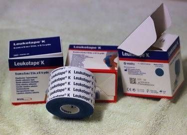 Drei Schachteln blaues Leukotape K Kinesiologie-Tape, wobei eine Rolle vorne liegt.