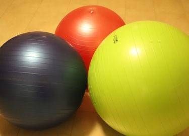 Ein violetter, ein roter und ein hellgrüner Gymnastikball liegen auf einem Holzboden.