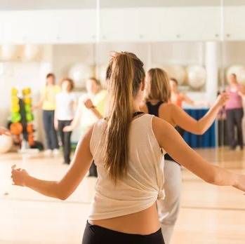 Menschen in einem hell erleuchteten Studio nehmen an einem Fitnesskurs teil; die Aufnahmen zeigen sie von hinten, während sie mit ausgestreckten Armen Übungen ausführen.