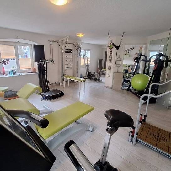 Ein heller Fitnessraum mit verschiedenen Trainingsgeräten, darunter eine Vibrationsplattform, eine limettengrüne Bank und ein Yogaball.