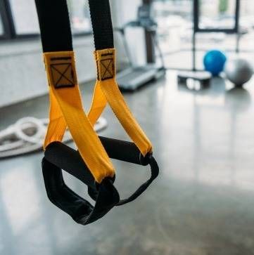 In einem Fitnessstudio hängen Schlingentrainingsgurte mit schwarzen Griffen und gelben Nylonbändern, im Hintergrund sind Trainingsgeräte zu sehen.