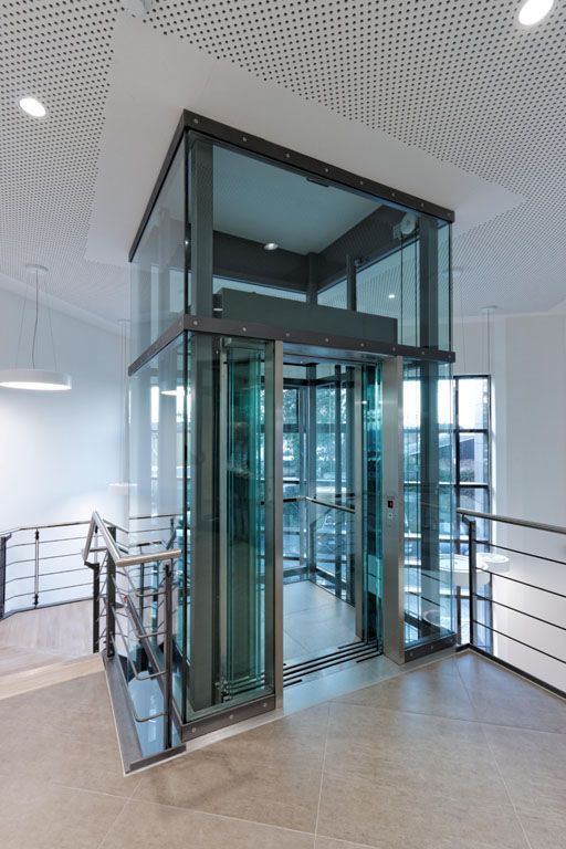 Ein gläserner Aufzug in einem Gebäude, zu dem eine Treppe hinaufführt.