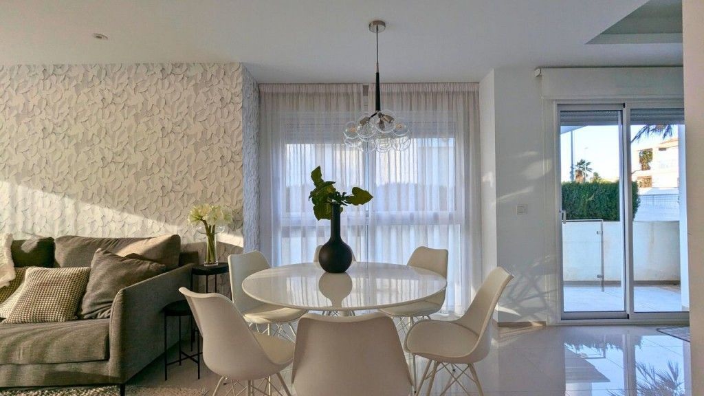 Comedor moderno con mesa y sillas blancas, cortinas transparentes y vista a un balcón.