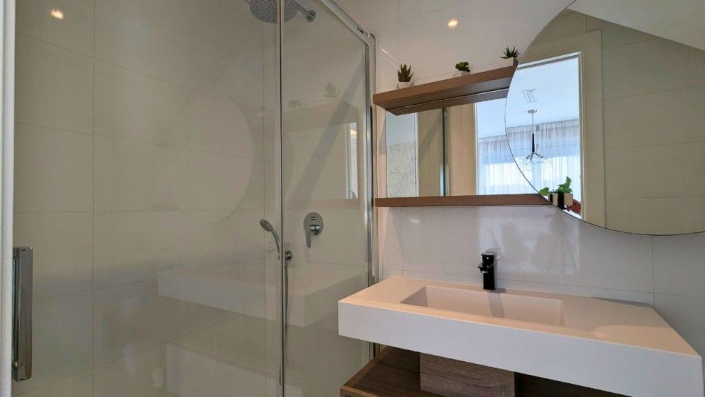 Baño con ducha de cristal, lavabo blanco y estantería de madera con pequeñas plantas.