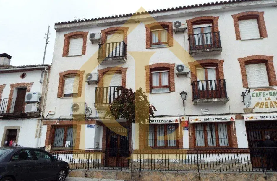 Edificio blanco de tres plantas con detalles rojos, balcones y tiendas en la planta baja. Hay un coche aparcado delante.