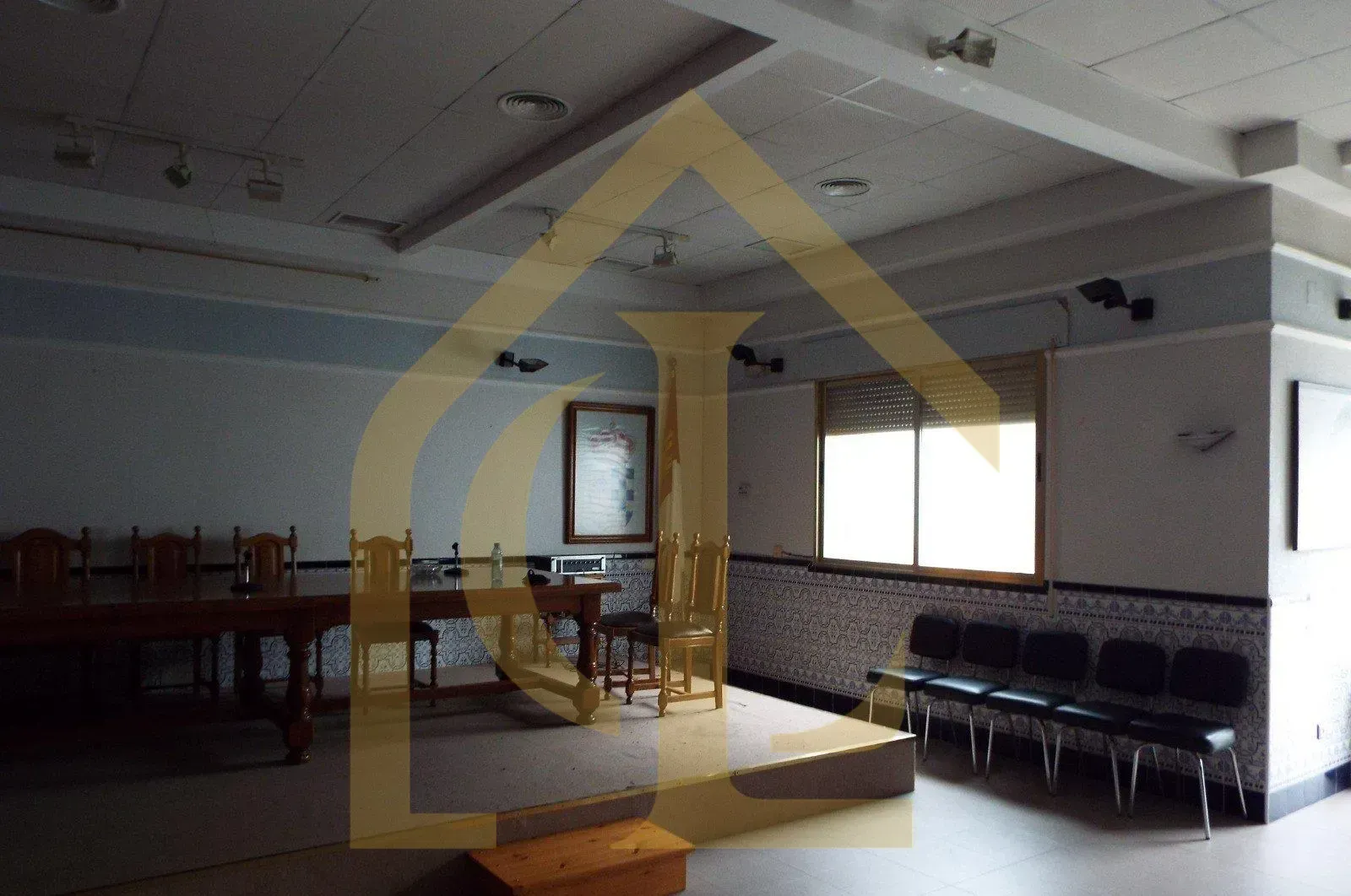 Sala vacía con mesas, sillas y escenario. Se ven una ventana y luminarias.