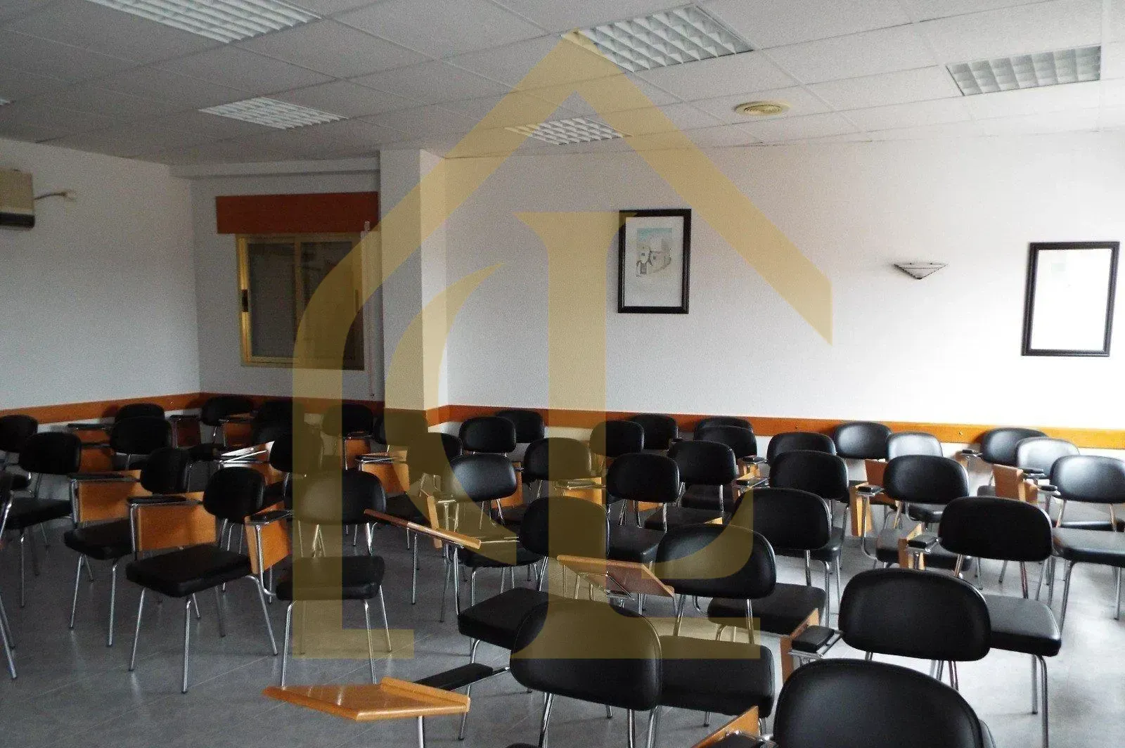 Aula con filas de sillas negras, algunas con pupitres incorporados. Paredes blancas, luces fluorescentes.