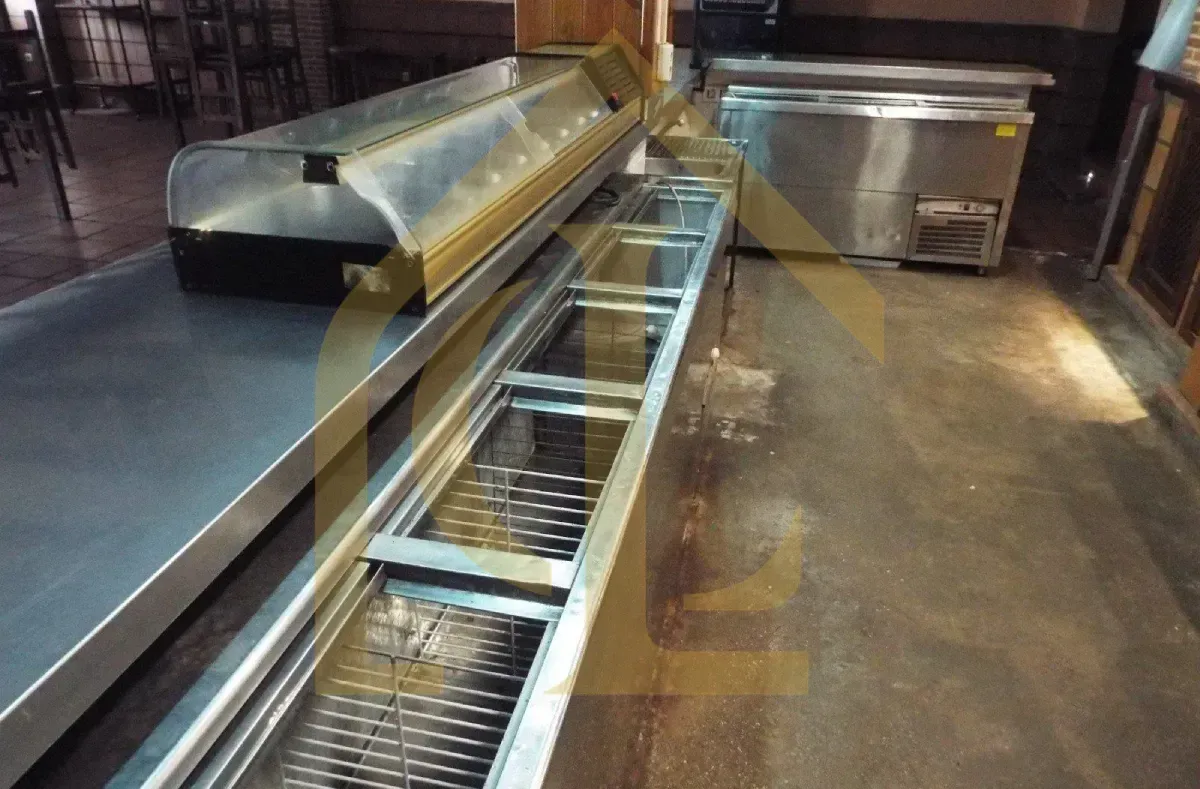 Equipamiento de cocina comercial de acero inoxidable; mostrador largo con barandillas y unidades de refrigeración.