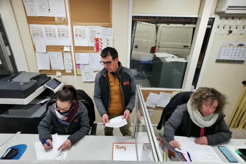 Tres personas trabajando en un escritorio, un hombre con papeles, otros escribiendo. Se ve una furgoneta al fondo a través de una ventana.