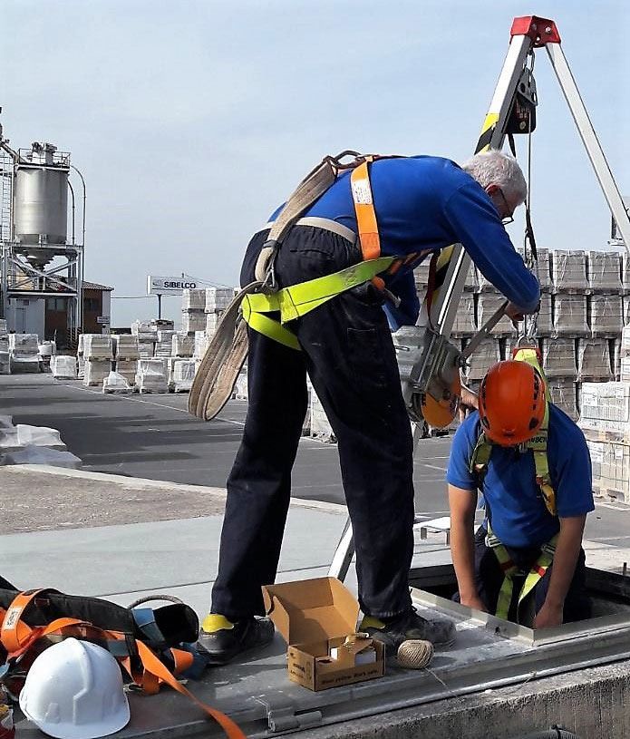 Dos trabajadores con arneses de seguridad en un tejado: uno de pie, trabajando en el equipo; el otro, arrodillado. Se ve una escalera con sistema de seguridad.