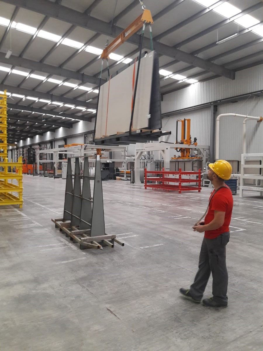 Un trabajador con casco opera una grúa levantando un gran panel de vidrio en una fábrica.
