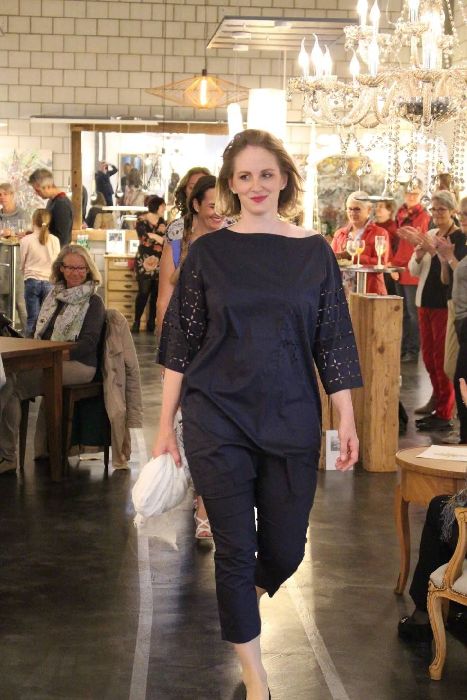 Eine Frau in marineblauem Outfit schreitet über einen Boutique-Laufsteg mit Kronleuchtern und sitzenden Gästen.