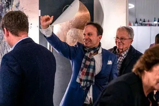 Ein Mann im blauen Anzug und Schal hebt bei einer Veranstaltung in einem geschlossenen Raum den Arm; andere Personen befinden sich in der Nähe.