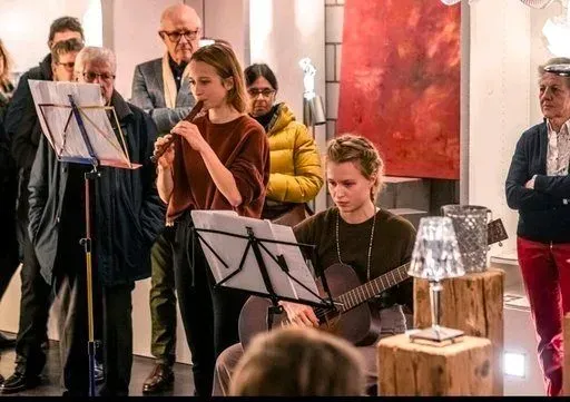 Eine kleine Gruppe spielt Musik in einem Innenraum; vor einer roten Wand stehen Notenständer und ein Gitarrist.