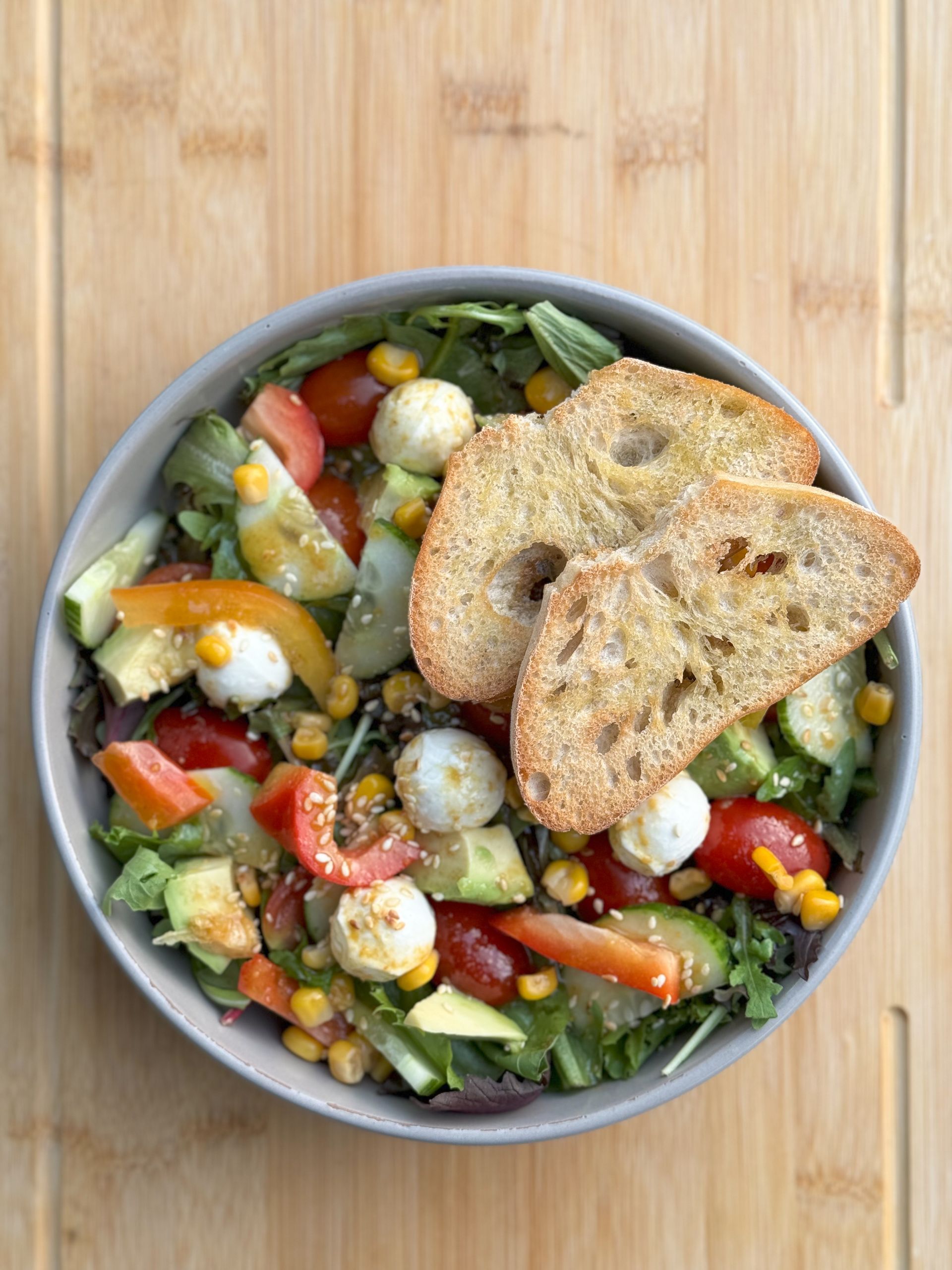 Eine Schüssel Salat mit Tomaten, Avocado, Mais und Croutons auf einem Holztisch.