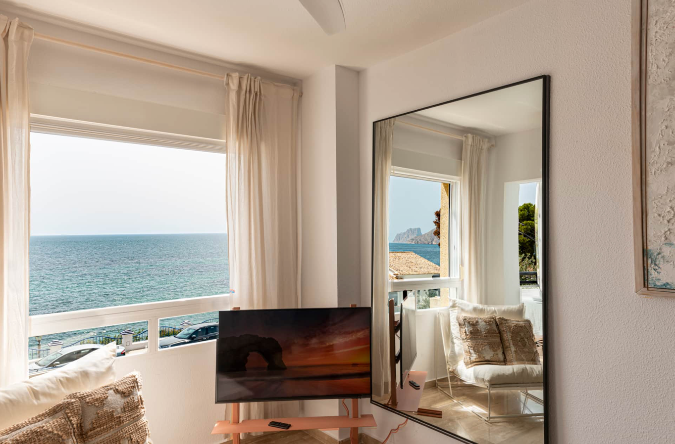 Room with a large window overlooking the ocean; a mirror reflects the view, and a TV shows a sunset scene.