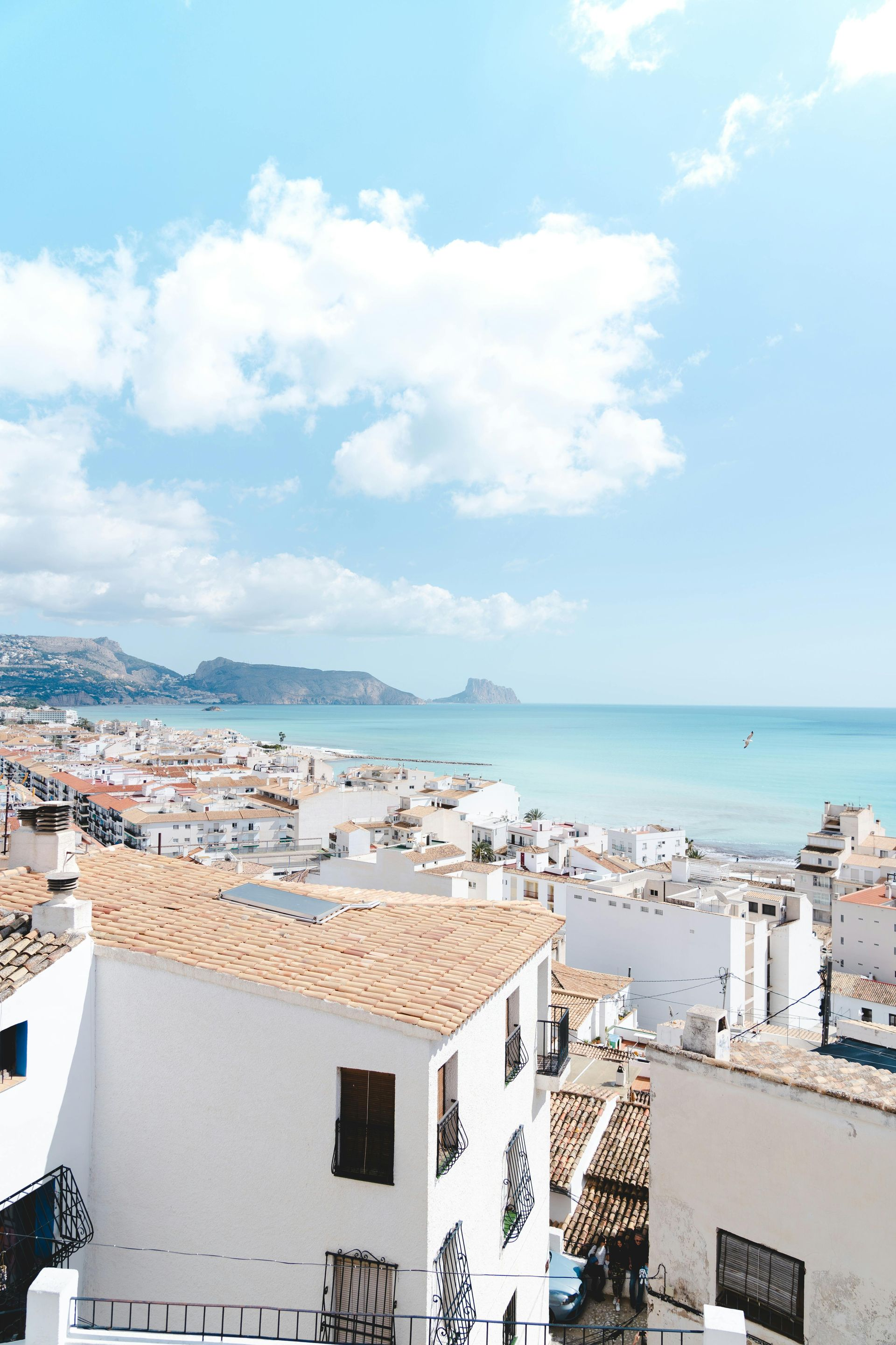 White buildings with terracotta roofs by a blue sea under a cloudy sky. Coastal town view.