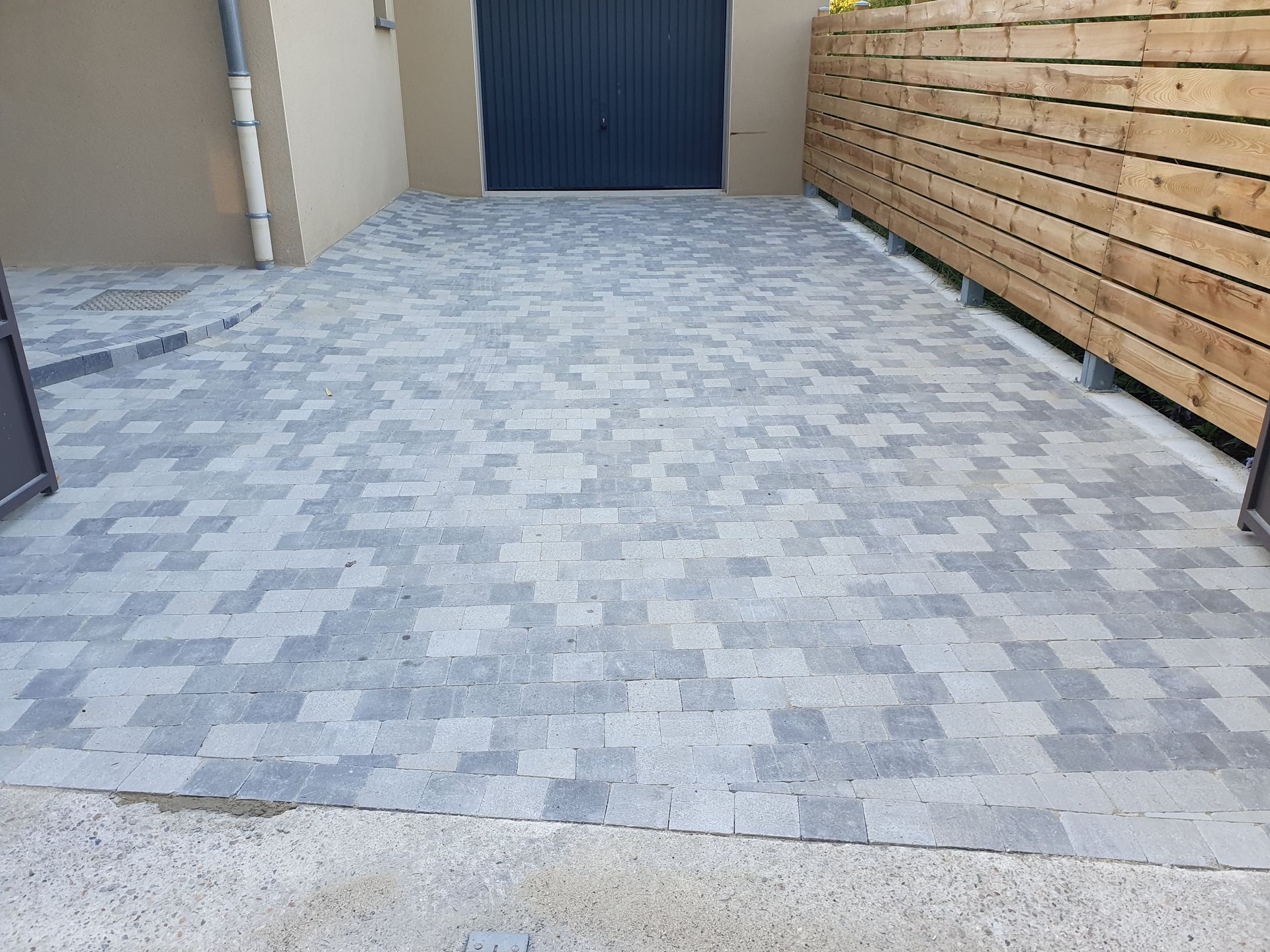 Allée pavée de briques grises menant à une porte de garage sombre, bordée d'une clôture en bois.