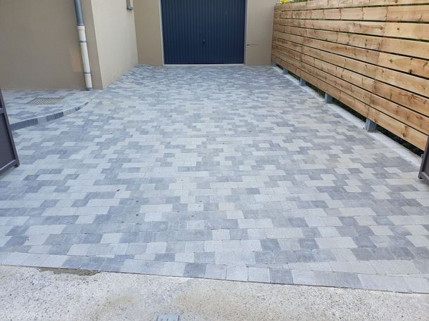 Allée pavée de briques grises menant à une porte de garage sombre, bordée d'une clôture en bois.
