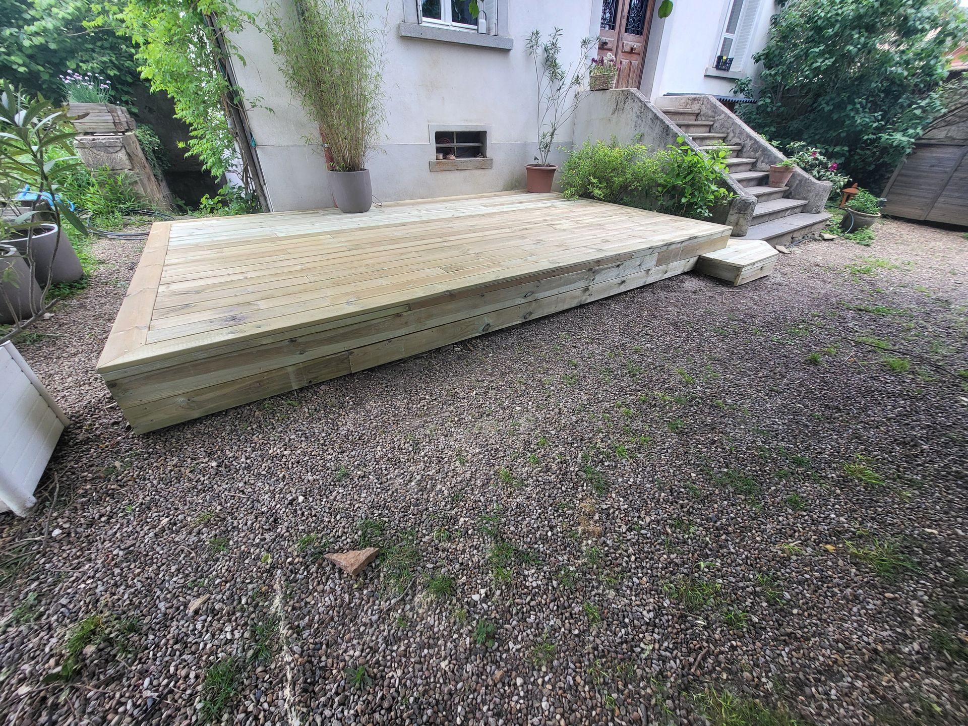 Terrasse en bois dans une cour gravillonnée, à côté d'un bâtiment avec un escalier et un bambou en pot.