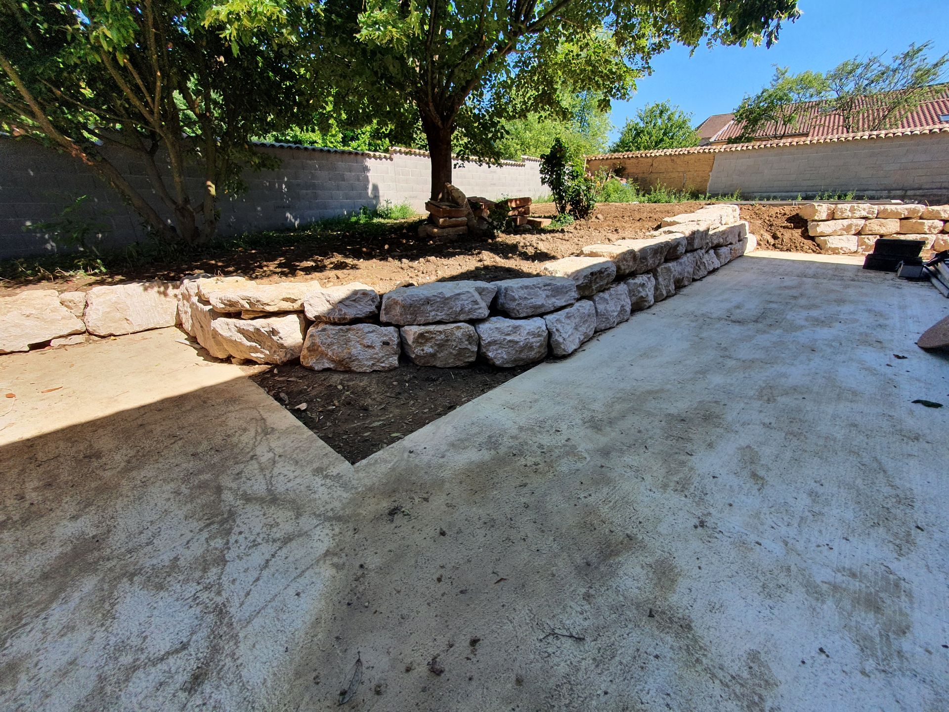 Mur de soutènement en pierre dans une cour arrière avec un arbre et une terrasse en béton.