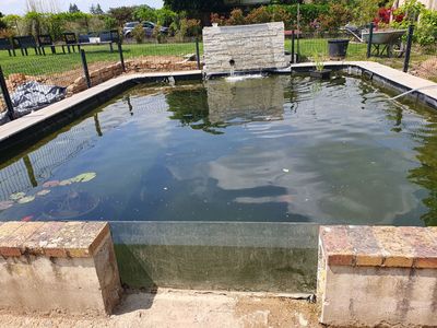 Bassin rectangulaire bordé de briques et agrémenté d'une fontaine en pierre, dans un jardin. L'eau reflète le ciel.